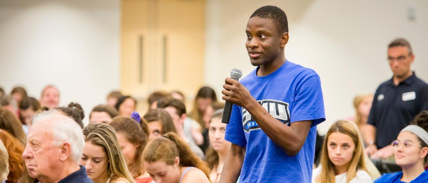 A student in a blue U N E t-shirt asks a question from the crowd of a President's Forum event